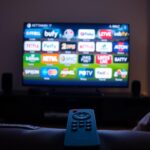 A cozy living room with a large 4K Smart TV screen glowing softly, displaying various sports channel logos and live footage. The room is dimly lit, creating a warm and inviting atmosphere. A sleek, modern remote control rests on a plush leather couch, hinting at the user's ability to easily navigate the IPTV service and access a wide range of live sports content. The scene conveys the excitement and convenience of watching sports events from the comfort of home with a top IPTV provider.